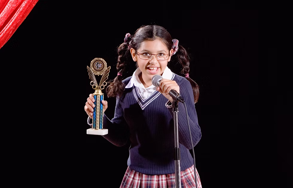 Young girl with pigtails and eyeglasses with her hand on a microphone and holding a trophy while she sings.